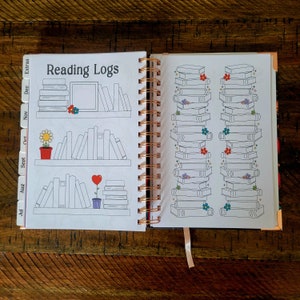 May include: A white reading log book with a pink, orange, and purple color scheme. The cover features the text "Reading Logs" and illustrations of bookshelves with books. The book has a spiral binding and a ribbon bookmark.