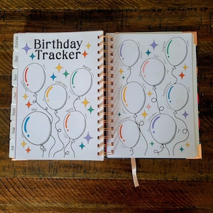 May include: A white planner with a colorful birthday tracker. The tracker features a design of balloons with each month of the year written on them. The planner has a spiral binding and a ribbon bookmark.
