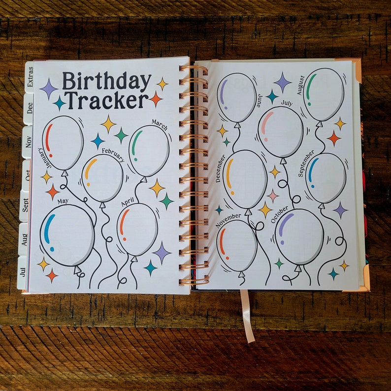 May include: A white planner with a colorful birthday tracker. The tracker features a design of balloons with each month of the year written on them. The planner has a spiral binding and a ribbon bookmark.