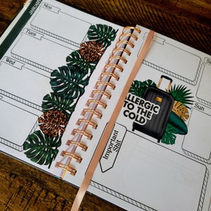 May include: A close-up of a planner with a gold spiral binding. The planner has a tropical leaf design and a sticker that says "Allergic to the Cold". The planner also has a sticker that says "Important Shit".