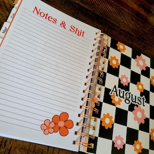 May include: A white planner with a black and white checkered design on the right side. The left side has lined pages with the text "Notes & Shit" at the top. The checkered side has the month "August" written in black text. The planner has a copper spiral binding.