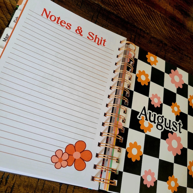 May include: A white planner with a black and white checkered design on the right side. The left side has lined pages with the text "Notes & Shit" at the top. The checkered side has the month "August" written in black text. The planner has a copper spiral binding.