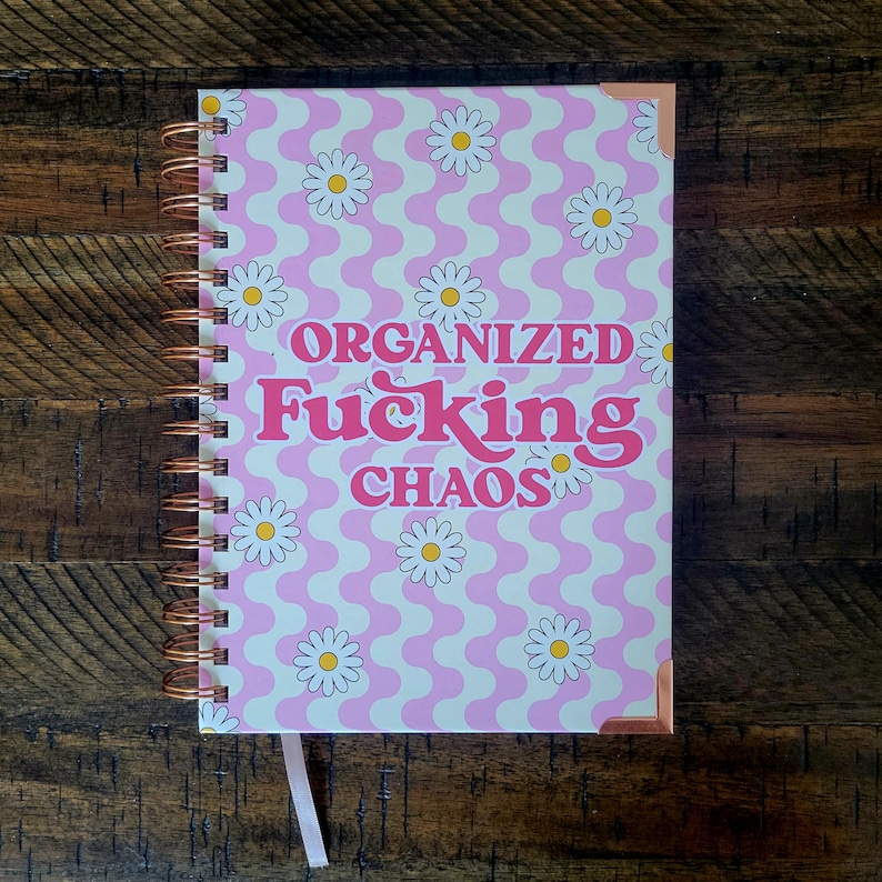 May include: A pink and white spiral bound notebook with a daisy pattern and the text "Organized Fucking Chaos" in pink and red letters.
