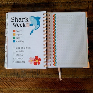 May include: A page from a shark week themed planner with a cartoon shark holding a coffee mug. The page has a calendar grid with boxes for each day of the month. The page also has a list of symptoms with checkboxes next to each one.