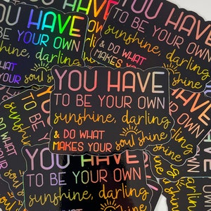 May include: A stack of black and rainbow holographic stickers with the text "You have to be your own sunshine, darling & do what makes your soul shine" repeated on each sticker.