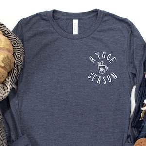 May include: A navy blue long-sleeved t-shirt with the text "Hygge Season" in white lettering. The text is in a circle with a stylized mug of hot chocolate with marshmallows and a cinnamon stick inside the circle.