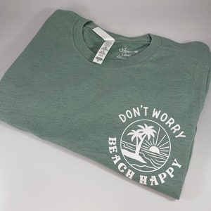 May include: A sage green t-shirt with a white circle graphic that says "Don't Worry Beach Happy" with a palm tree and sun graphic inside the circle.