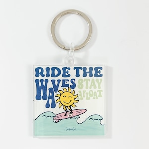 May include: A square keychain with a blue and yellow design. It features a smiling sun with a surfboard riding a wave. The text "Ride the Waves Stay Afloat" is written in blue and yellow letters.