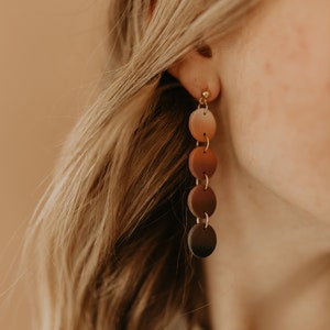 May include: A pair of long, dangle earrings with a gradient of brown tones. The earrings are made of round, flat discs that are connected by small gold rings. The discs are a light brown at the top and gradually get darker towards the bottom, ending in a dark brown color.