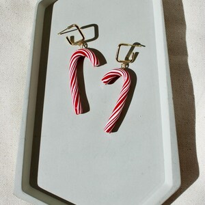 May include: A pair of festive candy cane earrings. Each earring features a red and white striped candy cane charm suspended from a gold-colored square hoop. The earrings are displayed on a light gray, hexagonal tray.
