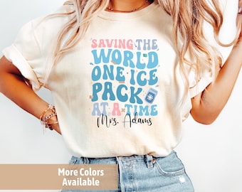 May include: A white t-shirt with a blue and pink graphic that says "Saving the World One Ice Pack at a Time" with the name "Mrs. Adams" underneath. The shirt is being worn by a person with long blonde hair and blue jeans.