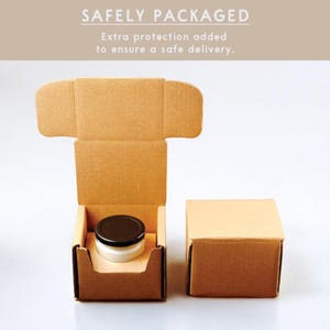 May include: A brown cardboard box with a lid is open to reveal a small white jar with a black lid. The jar is sitting inside a smaller brown cardboard box. The text "SAFELY PACKAGED" is printed on a white background above the boxes. The text below reads "Extra protection added to ensure a safe delivery."