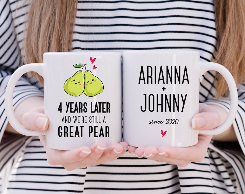 Puede incluir: Dos tazas blancas con texto negro y un dise&ntilde;o de pera. La taza de la izquierda dice "4 a&ntilde;os despu&eacute;s y seguimos siendo una gran pera". La taza de la derecha dice "Arianna + Johnny desde 2020".