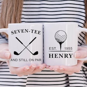 May include: Two white ceramic mugs with black text and graphics. One mug reads "SEVEN-TEE AND STILL ON PAR" with crossed golf clubs. The other mug features a golf ball on a tee and the name "HENRY" with "EST. 1955".