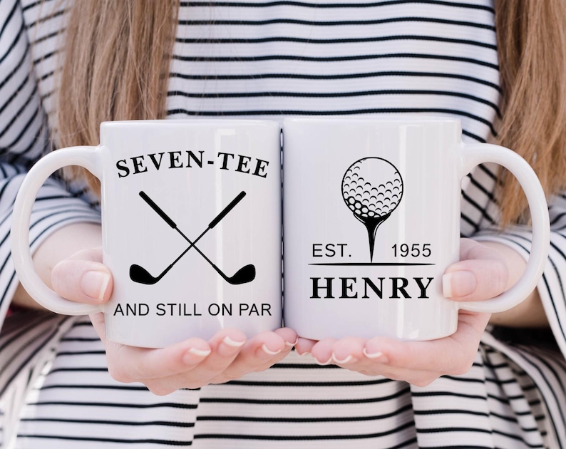 May include: Two white ceramic mugs with black text and graphics. One mug reads "SEVEN-TEE AND STILL ON PAR" with crossed golf clubs. The other mug features a golf ball on a tee and the name "HENRY" with "EST. 1955".