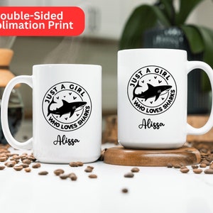 May include: Two white ceramic mugs with a black shark graphic and the text "JUST A GIRL WHO LOVES SHARKS" and the name "Alissa". The mugs are on a wooden coaster and a surface scattered with coffee beans. The top left corner has a red banner that says "Double-Sided Sublimation Print".