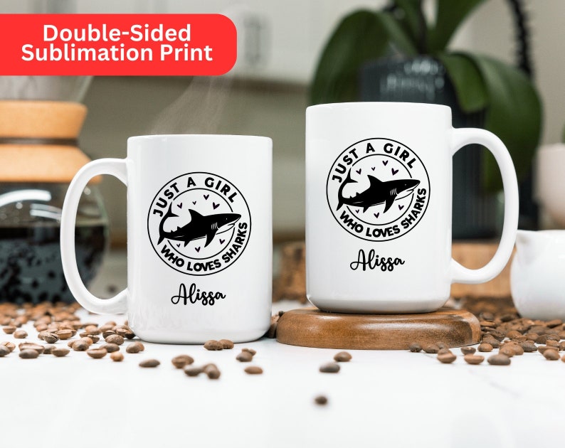May include: Two white ceramic mugs with a black shark graphic and the text "JUST A GIRL WHO LOVES SHARKS" and the name "Alissa". The mugs are on a wooden coaster and a surface scattered with coffee beans. The top left corner has a red banner that says "Double-Sided Sublimation Print".