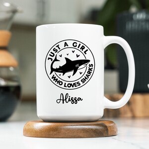 May include: White ceramic mug with a black shark graphic and the text "JUST A GIRL WHO LOVES SHARKS" in a circular design. The name "Alissa" is written below the graphic. The mug is on a wooden coaster.