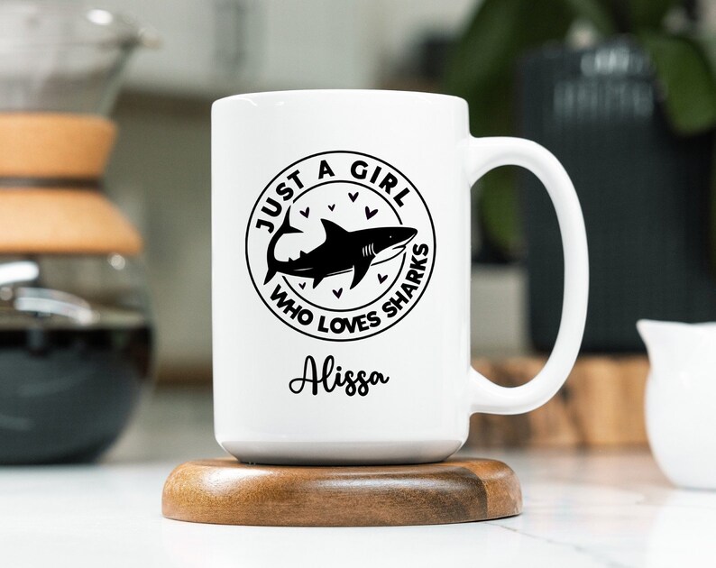May include: White ceramic mug with a black shark graphic and the text "JUST A GIRL WHO LOVES SHARKS" in a circular design. The name "Alissa" is written below the graphic. The mug is on a wooden coaster.