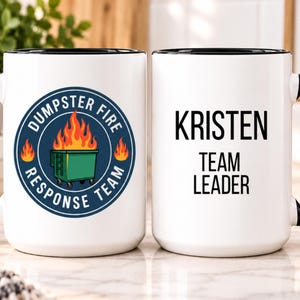May include: Two white ceramic mugs with black handles and rims. One mug features a circular blue emblem with a green dumpster on fire and the text DUMPSTER FIRE RESPONSE TEAM. The other mug displays the black text KRISTEN TEAM LEADER.
