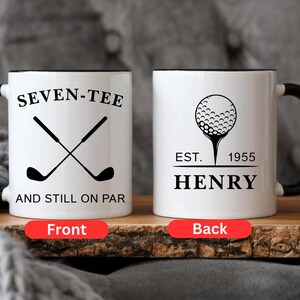May include: White ceramic mug with a black handle and rim. The front of the mug features the text "SEVEN-TEE" above crossed golf clubs and "AND STILL ON PAR". The back has a golf ball graphic, the text "EST. 1955 HENRY".