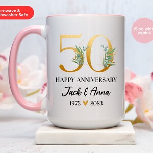 May include: White ceramic mug with a pink handle. The mug has a gold number 50 with green floral accents. The text "Happy Anniversary Jack & Anna 1973 2023" is printed below the number. The mug is 15 ounces and is white with a pink rim.
