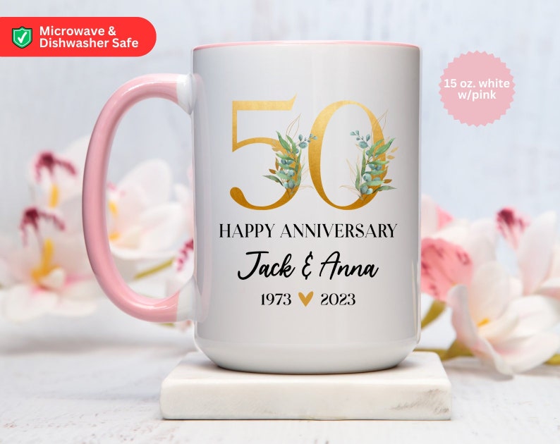 May include: White ceramic mug with a pink handle. The mug has a gold number 50 with green floral accents. The text "Happy Anniversary Jack & Anna 1973 2023" is printed below the number. The mug is 15 ounces and is white with a pink rim.