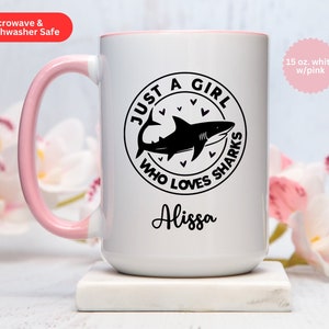 May include: White 15 oz. ceramic mug with a pink handle and rim. The mug features a black graphic of a shark inside a circle with the text "JUST A GIRL WHO LOVES SHARKS" and the name "Alissa". The mug is microwave and dishwasher safe.