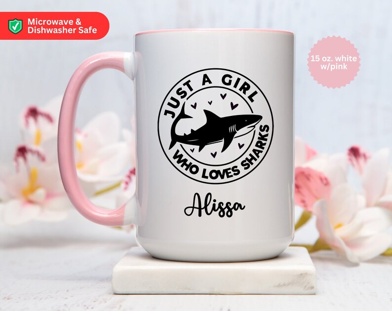 May include: White 15 oz. ceramic mug with a pink handle and rim. The mug features a black graphic of a shark inside a circle with the text "JUST A GIRL WHO LOVES SHARKS" and the name "Alissa". The mug is microwave and dishwasher safe.