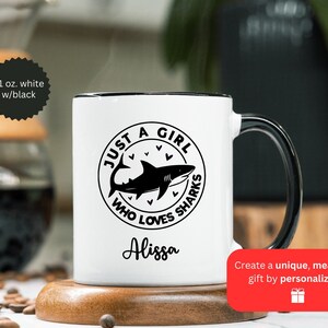May include: White ceramic mug with a black handle and rim. The mug features a black graphic of a shark inside a circle with the text "JUST A GIRL WHO LOVES SHARKS" and the name "Alissa". The mug is 11 oz.