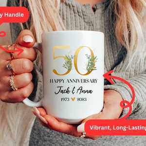 May include: White ceramic mug with a gold 50th anniversary design, featuring a floral motif and the text "Happy Anniversary Jack & Anna 1973 &hearts; 2023".