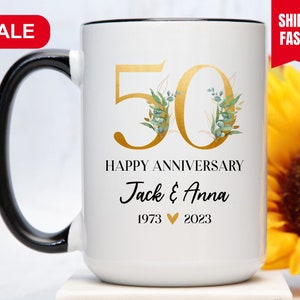 May include: White ceramic mug with a black handle. The mug has a gold 50 with green and gold floral accents. The text "Happy Anniversary Jack & Anna 1973 &hearts; 2023" is printed on the mug.