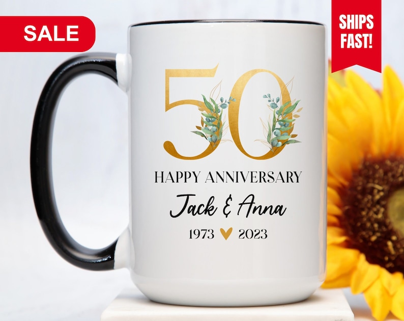 May include: White ceramic mug with a black handle. The mug has a gold 50 with green and gold floral accents. The text "Happy Anniversary Jack & Anna 1973 &hearts; 2023" is printed on the mug.