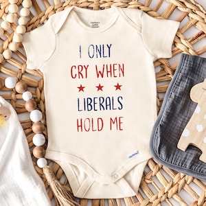 May include: A white baby onesie with the text "I only cry when liberals hold me" in red and blue lettering. There are three red stars above the word "liberals".