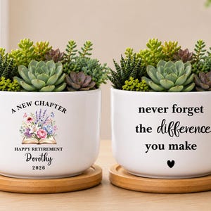 May include: Two white ceramic planters on bamboo saucers, each filled with assorted green succulents. The left planter features a floral book graphic with the text A NEW CHAPTER HAPPY RETIREMENT Dorothy 2026. The right planter reads never forget the difference you make with a heart icon.