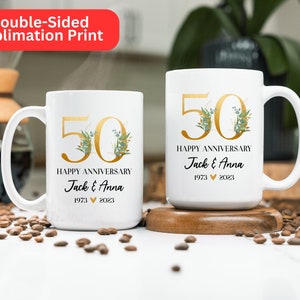 May include: Two white ceramic mugs with gold lettering and green floral accents. The mugs are personalized with the text "50 Happy Anniversary Jack & Anna 1973 2023".