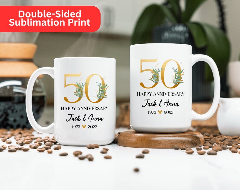 May include: Two white ceramic mugs with gold lettering and green floral accents. The mugs are personalized with the text "50 Happy Anniversary Jack & Anna 1973 2023".