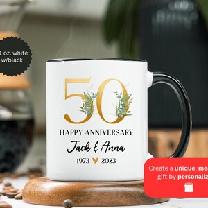 May include: White ceramic mug with a black rim and handle. The mug features a gold number 50 with green floral accents and the text "Happy Anniversary Jack & Anna 1973 2023".