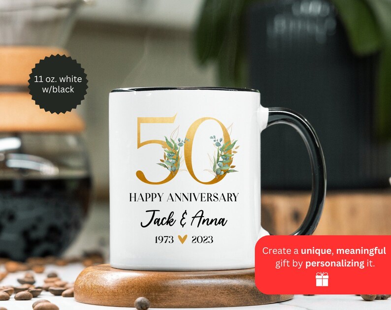 May include: White ceramic mug with a black rim and handle. The mug features a gold number 50 with green floral accents and the text "Happy Anniversary Jack & Anna 1973 2023".