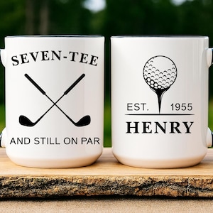 May include: White ceramic mug with a black handle and rim. The mug features golf-themed text and graphics, including crossed golf clubs and a golf ball on a tee. The text reads "SEVEN-TEE AND STILL ON PAR" and "EST. 1955 HENRY".
