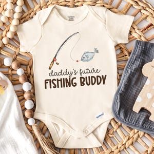 May include: Cream-colored baby onesie with the text "daddy's future FISHING BUDDY" and a fishing rod graphic. A gray bib and wooden giraffe toy are next to the onesie. The onesie is made of soft material.