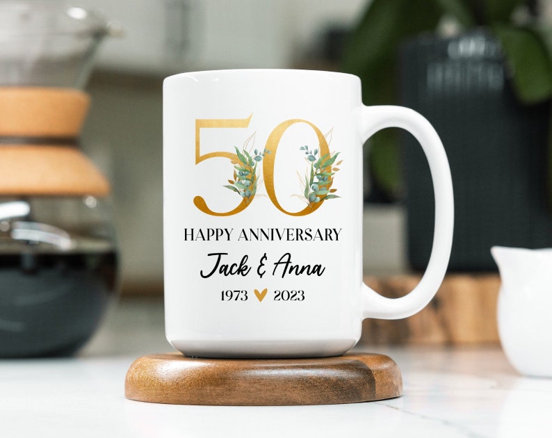 May include: White ceramic mug with a gold 50 and floral design. The mug says "Happy Anniversary Jack & Anna 1973 &hearts; 2023".