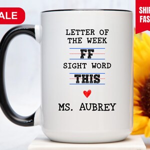 May include: White ceramic coffee mug with a black handle. The mug has a black rim and features the text "Letter of the Week" with the letter "F" written twice on a lined paper design. The text "Sight Word" is written below the letter "F" and the word "This" is written below that. A red heart is below the word "This" and the text "Ms. Aubrey" is written below the heart.