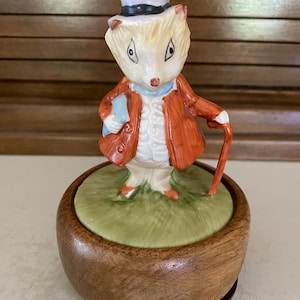 May include: A porcelain figurine of a hedgehog wearing a top hat and a red jacket, holding a walking stick. The figurine is on a wooden music box base.