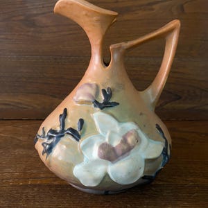 May include: A brown ceramic pitcher with a white magnolia flower design. The pitcher has a unique, curved handle and a spout.