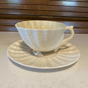 May include: A cream-colored teacup and saucer set. The cup has a unique shell-like design with a handle and sits on three small feet. The saucer mirrors the cup's design, creating a cohesive and elegant look. The set is made of ceramic.