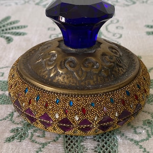 May include: A round, gold-colored trinket box with a blue glass knob. The box is decorated with small, gold beads and purple and red gemstones.