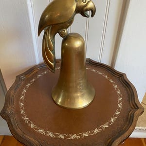 May include: A brass parrot figurine perched atop a bell-shaped base. The parrot is detailed with feathers and has a realistic beak. The bell is smooth and polished.