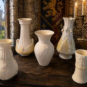 May include: A collection of five white ceramic vases with varied textures and shapes, including fluted rims, embossed floral patterns, and leaf motifs, arranged on a dark wooden surface with a glossy finish and scalloped edges.