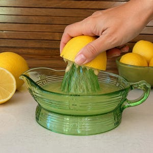 May include: A vintage green depression glass citrus juicer with a ribbed exterior design and a handle. The juicer features a central reamer cone and a wide basin for collecting juice, accompanied by several bright yellow lemons nearby.